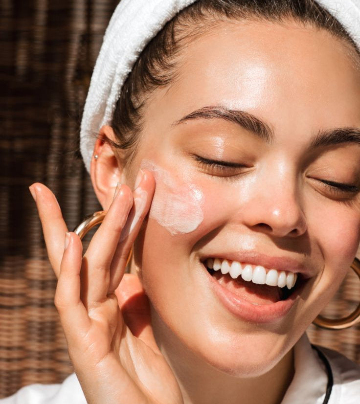 Woman applying cream to her face with a towel on her head, smiling outdoors.